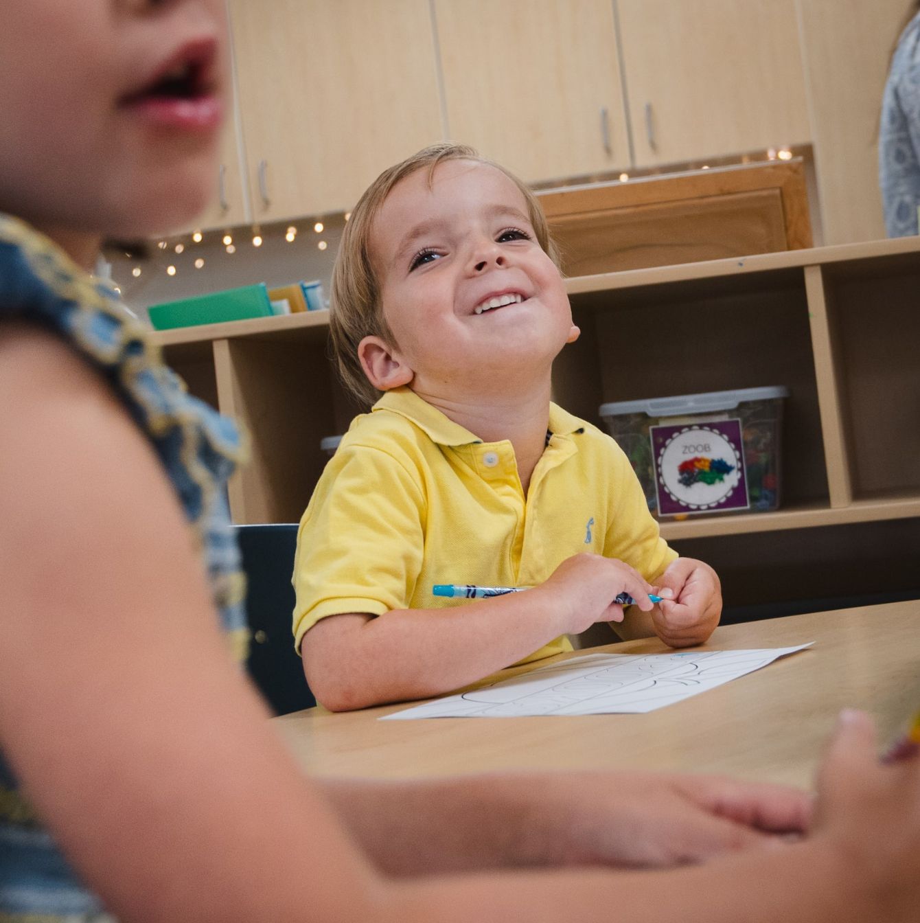 Smiling young boy in a yellow shirt enjoying coloring with crayons at a classroom table, with other children and classroom supplies in the background.